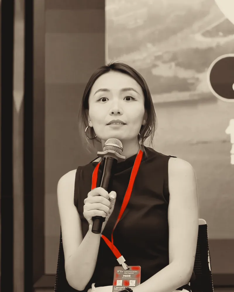 A speaker with a microphone, a red lanyard across her chest, in front of the summit stage screen.